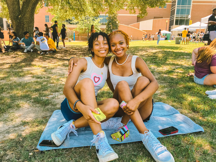 Two female students sitting on a towel playing cards outside. 