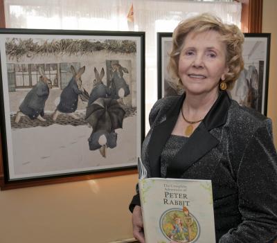 Janette Kennedy Tibbitts stands next to two paintings featuring the work of famed children's literature artist Beatrix Potter, creator of the “Peter Rabbit” character, while holding a copy of a book about the famed rabbit.