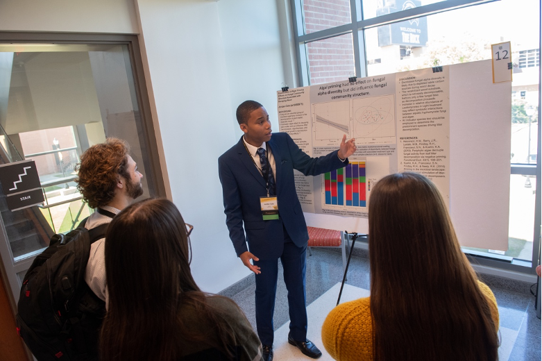 Forensics (Biological Sciences) major Jordan Cole received a 2022 Drapeau Summer Research Grant to study the effects of algae and fungi on wood decay under the mentorship of Biological Sciences professor, Dr. Kevin Kuehn. Here he is shown explaining his DCUR-funded research project to UGS 2023 attendees. 
